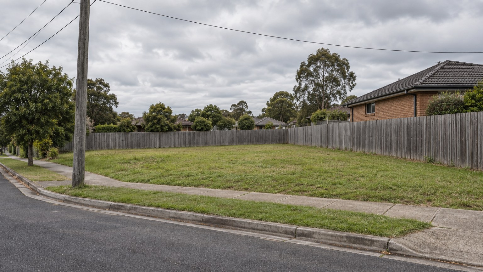 Illustrative view of an empty grassed suburban corner lot with a timber paling fence and a street power pole carrying overhead wires at the front boundary