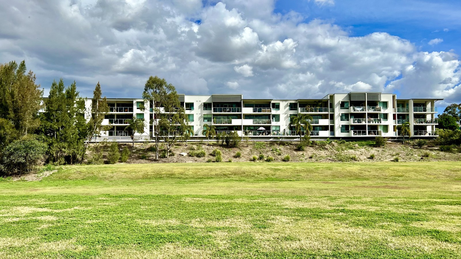 A long four-storey apartment building with white walls and glazed balconies set on a rise above a wide green park, under a cloudy blue sky